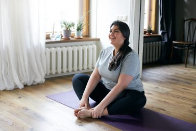 Cheerful attractive young overweight woman in activewear choosing healthy lifestyle, sitting on mat with hands on bare feet, doing butterfly yoga exercise, stretching thighs. Body shape and activity