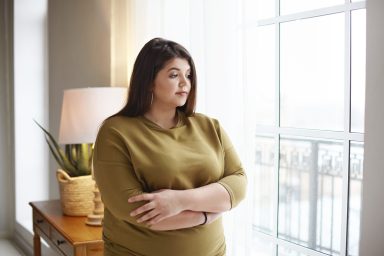 People, lifestyle and body positivity concept. Beautiful young brunette female with extra pounds standing by window at home, crossing arms on her chest, having thoughtful pensive look, pondering