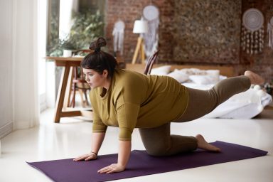 Sports, activity, fitness and weight loss concept. Indoor image of concentrated self determined young plus size woman in leggings and t-shirt exercising on mat, lifting one leg, trying to hold balance