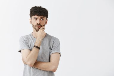 Waist-up shot of creative and artistic caucasian male model with beard holding hand on jaw and chin gazing right with spaced out expression thinking making plan over gray background. Body language concept